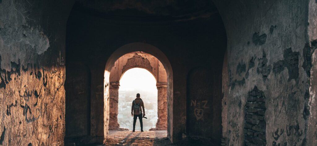 A man standing in an old, weathered archway with a backpack, overlooking a cityscape at sunrise or sunset.