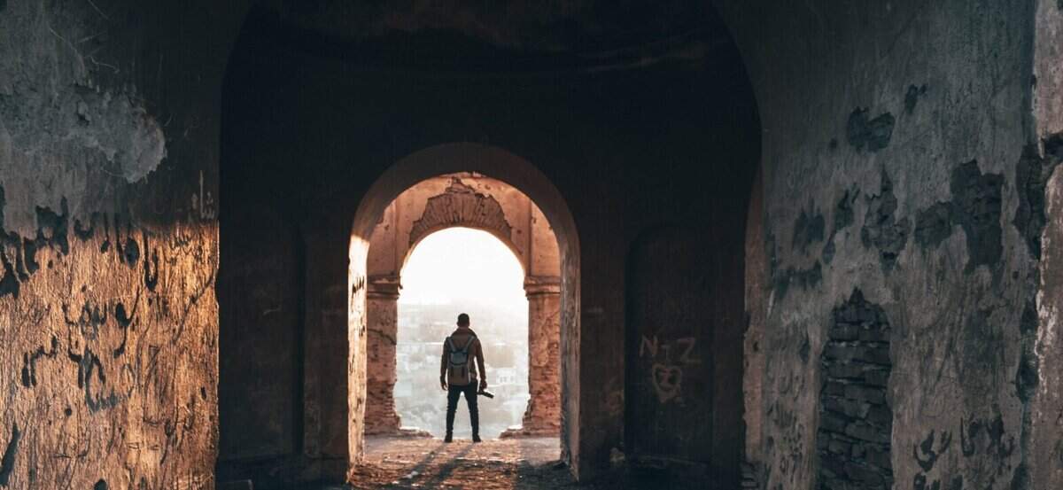 A man standing in an old, weathered archway with a backpack, overlooking a cityscape at sunrise or sunset.