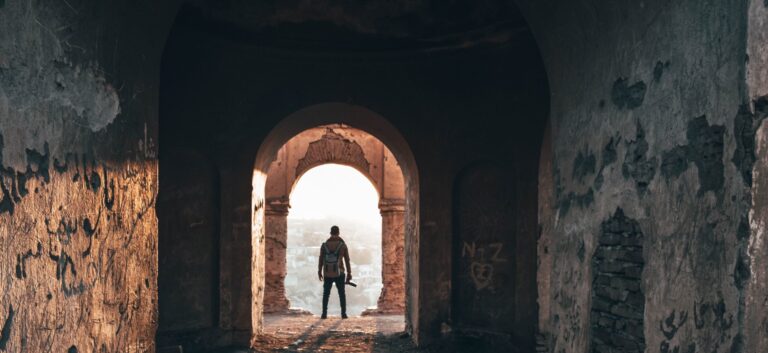 A man standing in an old, weathered archway with a backpack, overlooking a cityscape at sunrise or sunset.