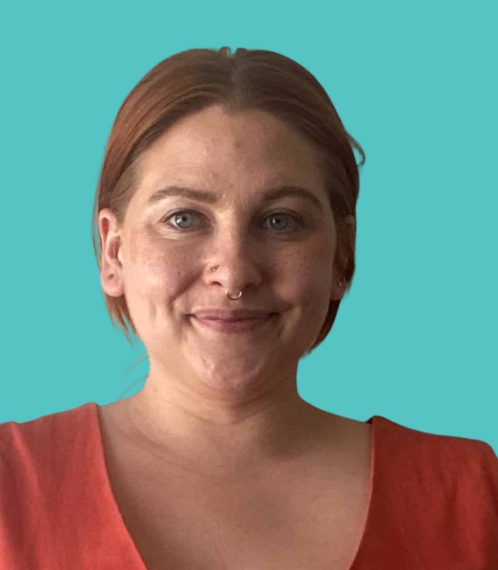 Confident woman with short red hair and septum piercing, smiling in front of a teal background.